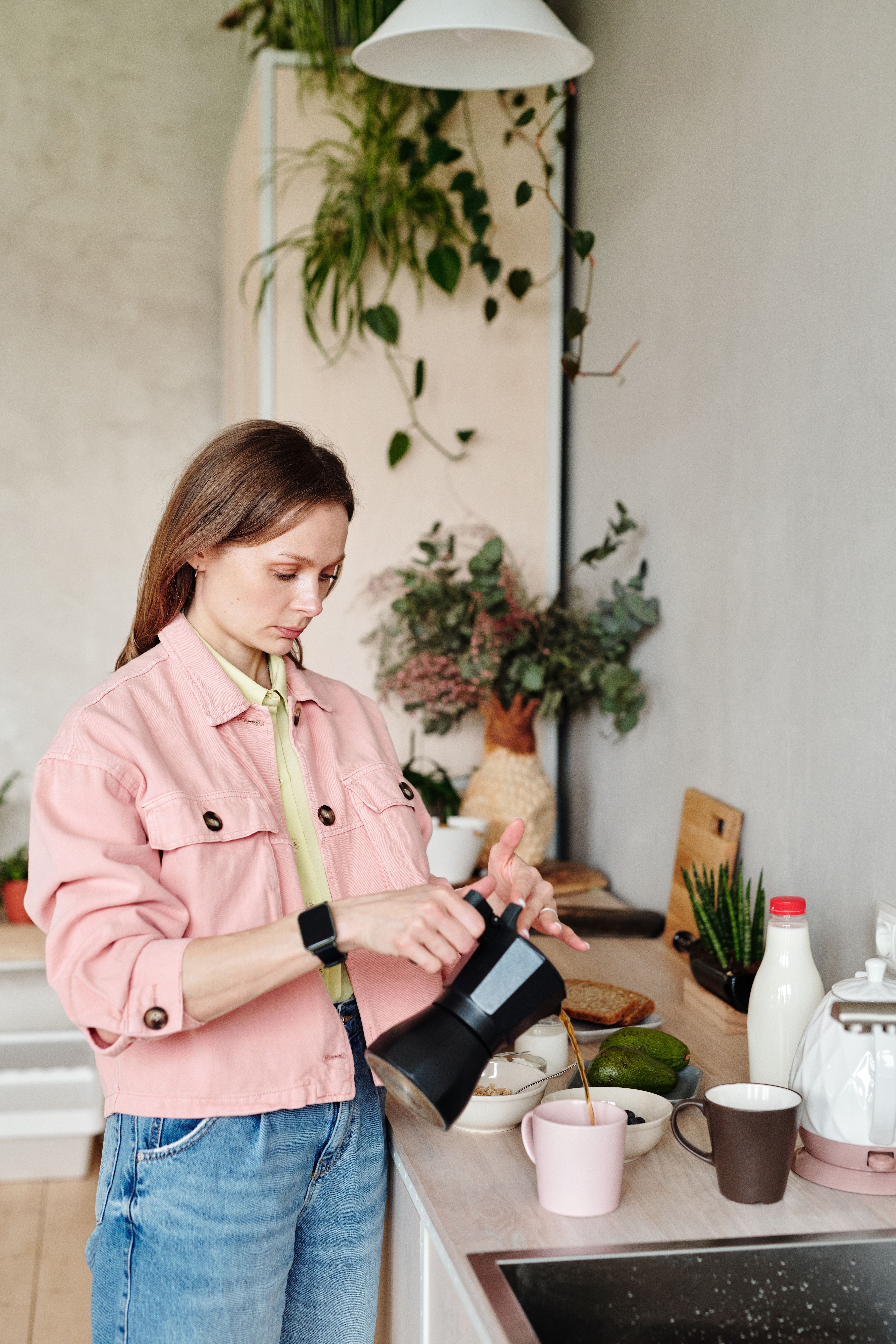 A woman in a pink jacket pours coffee in a bright, modern kitchen surrounded by plants and kitchenware.