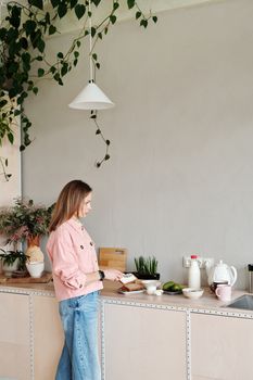 Woman slicing bread on kitchen counter surrounded by plants. Bright, indoor setting.