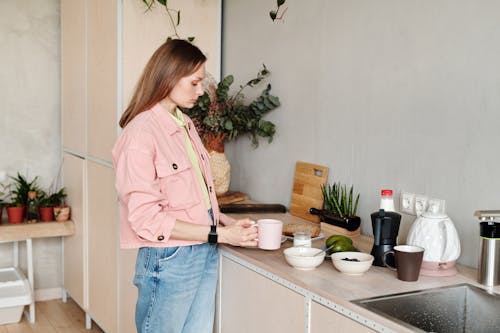 Woman in Pink Button Up Shirt and Blue Denim Jeans Standing Near Kitchen Sink