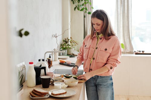 Woman in Pink Button Up Shirt and Blue Denim Jeans Holding Sliced Avocado