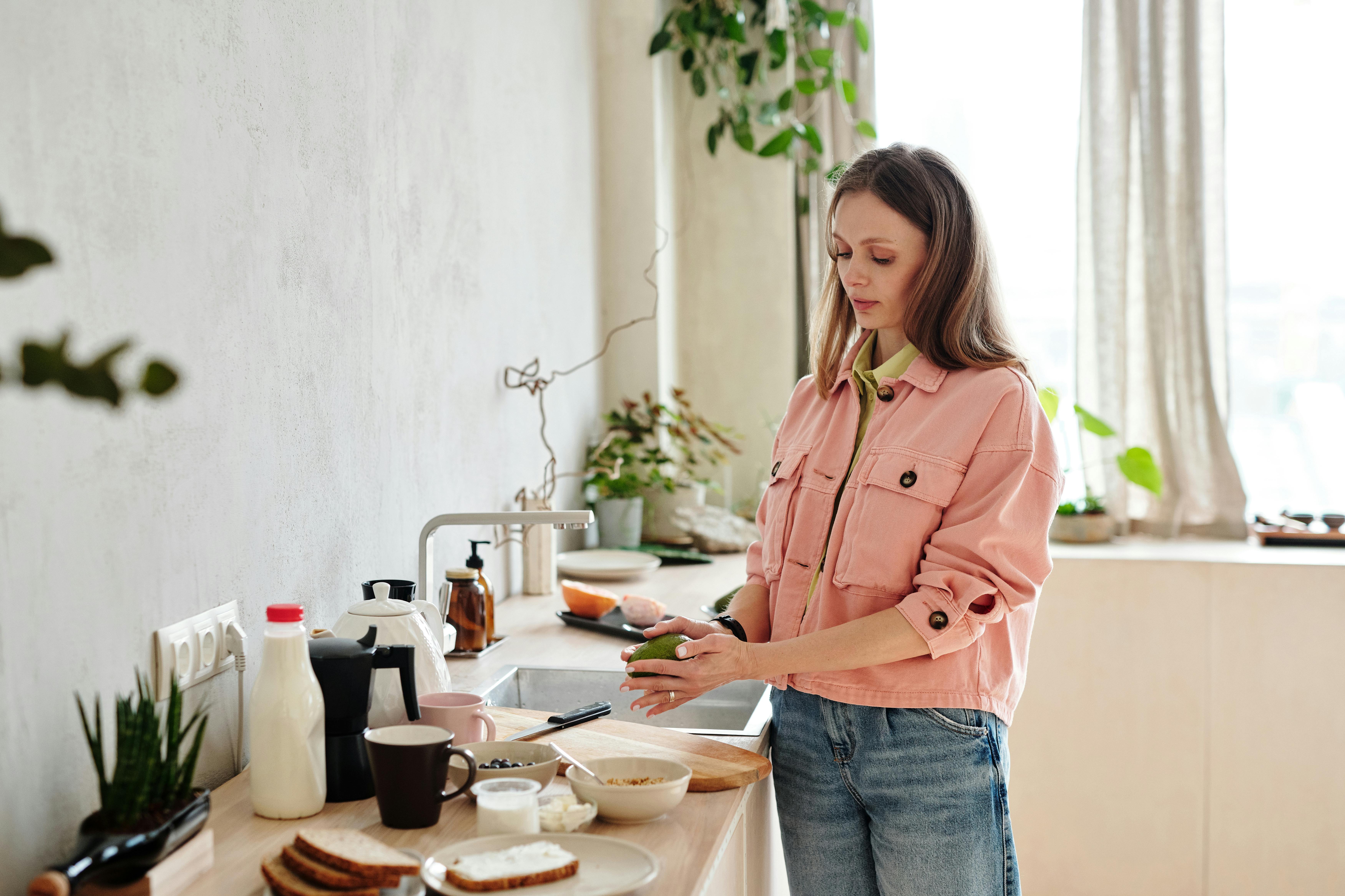 Young woman in pink jacket preparing avocado toast in a sunlit kitchen with houseplants.
