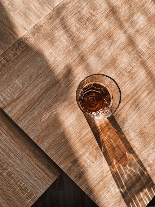 From above of transparent glass with beverage placed on wooden table and illuminated by sunlight
