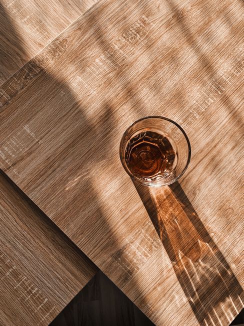 From above of transparent glass with beverage placed on wooden table and illuminated by sunlight