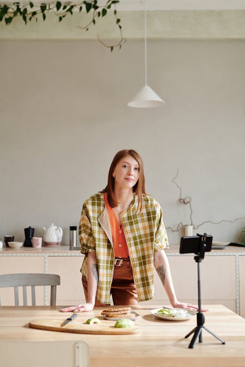 Woman in Green and Orange Plaid Shirt Standing beside Brown Wooden Table Vlogging