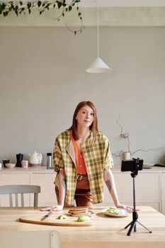 Woman vlogging food preparation in cozy kitchen with smartphone and tripod.