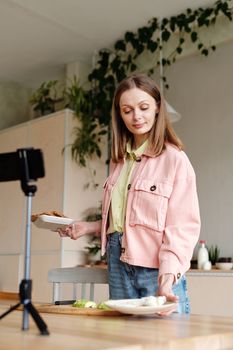 Woman in a pink jacket preparing food while vlogging in a stylish kitchen.