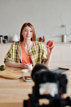 Young woman holding a grapefruit while filming a kitchen vlog at home.