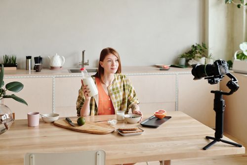 Free A woman filming a vlogging session in a modern kitchen with food items and camera setup. Indoor setting. Stock Photo