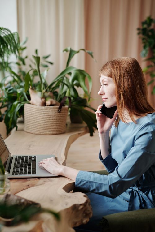 Free Caucasian woman in blue dress multitasking with laptop and smartphone at home office setting. Stock Photo