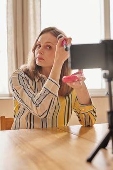 Caucasian woman using a makeup brush while recording a beauty vlog indoors.