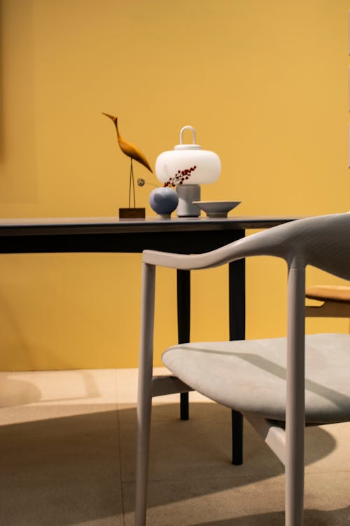 Contemporary style flat with lamp near small vase with twig and wooden statuette of bird on desk between chair and yellow wall