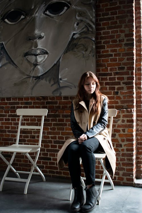 Young wistful female in coat and boots looking down while sitting on wooden stool in loft style building with picture on wall and concrete floor