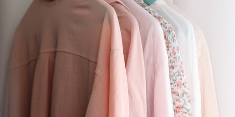 Modern shirts of different pink color shades on plastic hangers with metal hooks hanging on wooden beam in wardrobe near white concrete wall in apartment