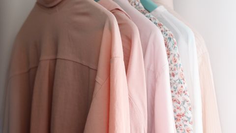 Modern shirts of different pink color shades on plastic hangers with metal hooks hanging on wooden beam in wardrobe near white concrete wall in apartment