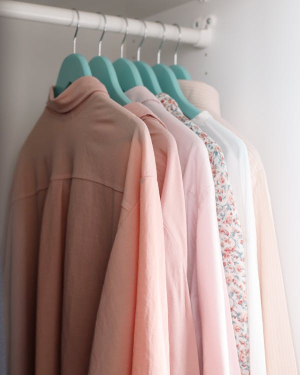 Modern shirts of different pink color shades on plastic hangers with metal hooks hanging on wooden beam in wardrobe near white concrete wall in apartment