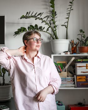 Plump contemplative female in casual shirt and eyewear with blushing cheeks touching neck and looking away while standing behind shelf with stationery and green plant in apartment
