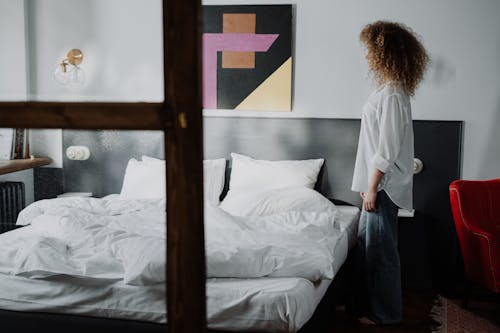 Free Woman in White Long Sleeve Shirt and Blue Denim Jeans Standing on Bed Stock Photo