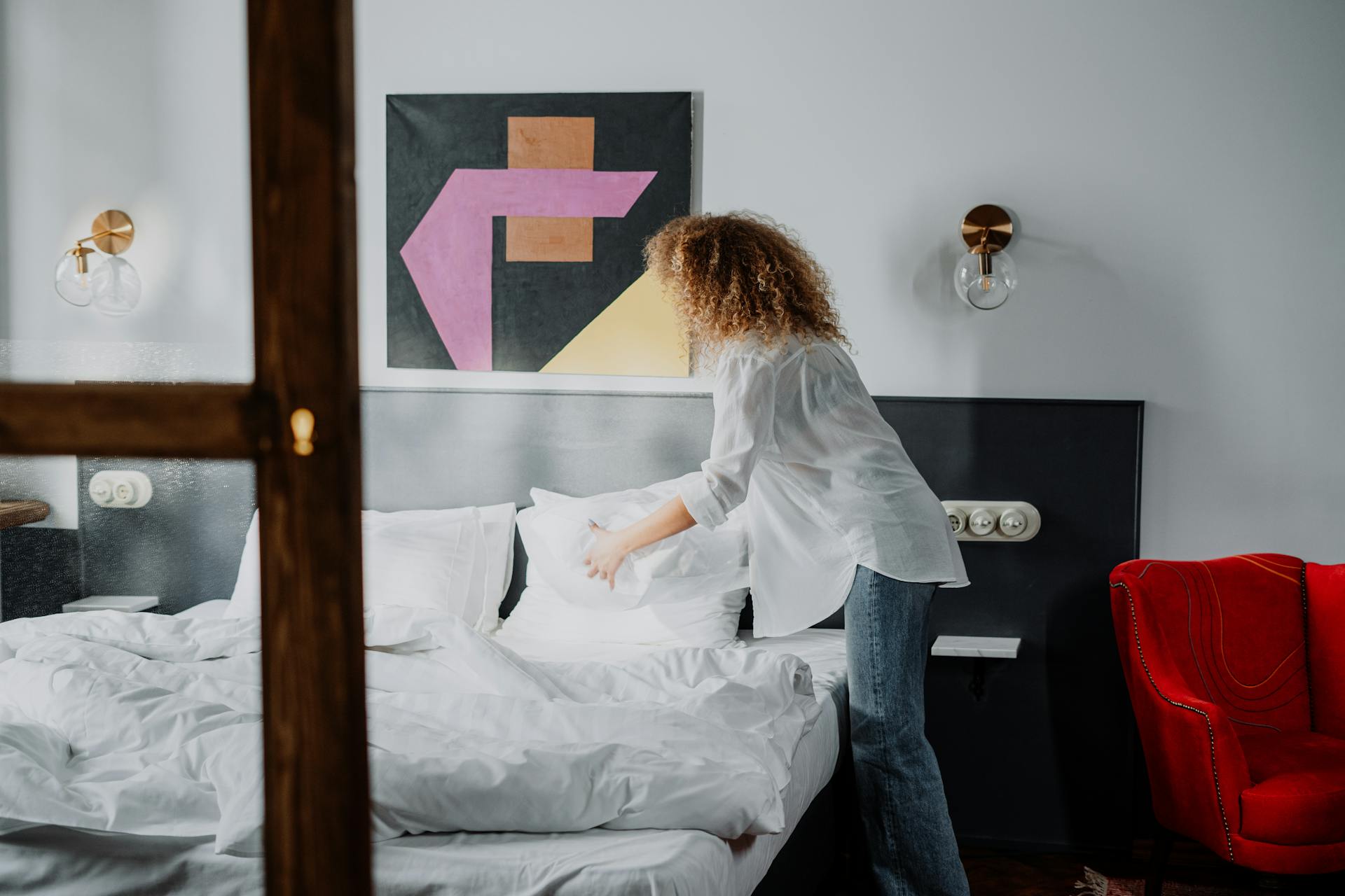 Woman adjusting pillows in a stylish bedroom with modern decor and bright lighting.