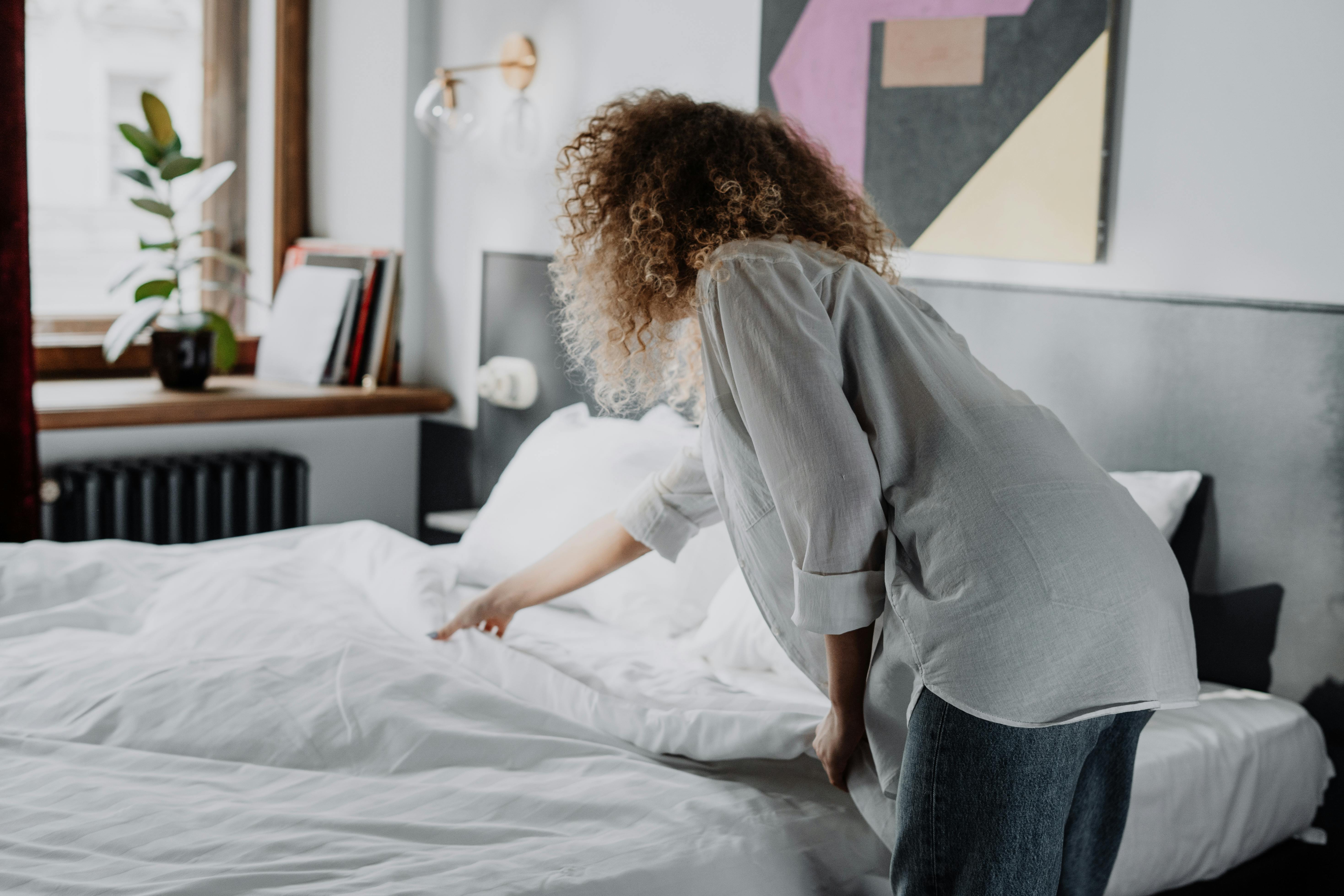 Free A woman with curly hair making a bed in a modern, cozy bedroom with natural light. Stock Photo