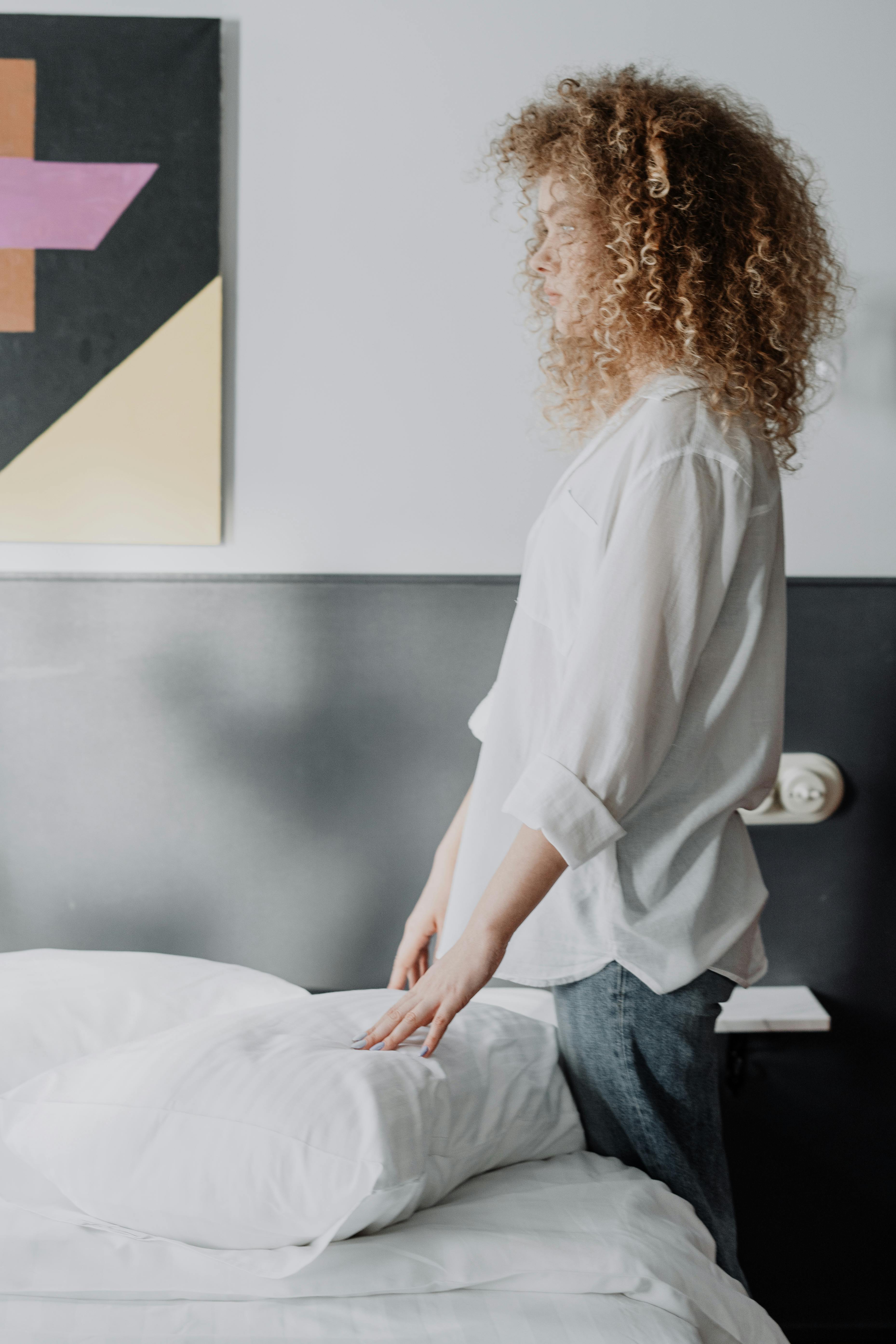 A woman with curly hair tidies up bed linens in a contemporary bedroom setting.
