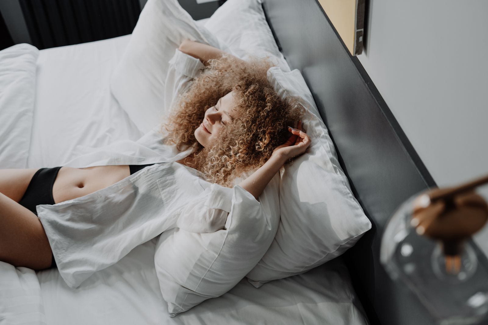 Woman with curly hair smiling and stretching on white pillows in a bed lit by warm sunlight