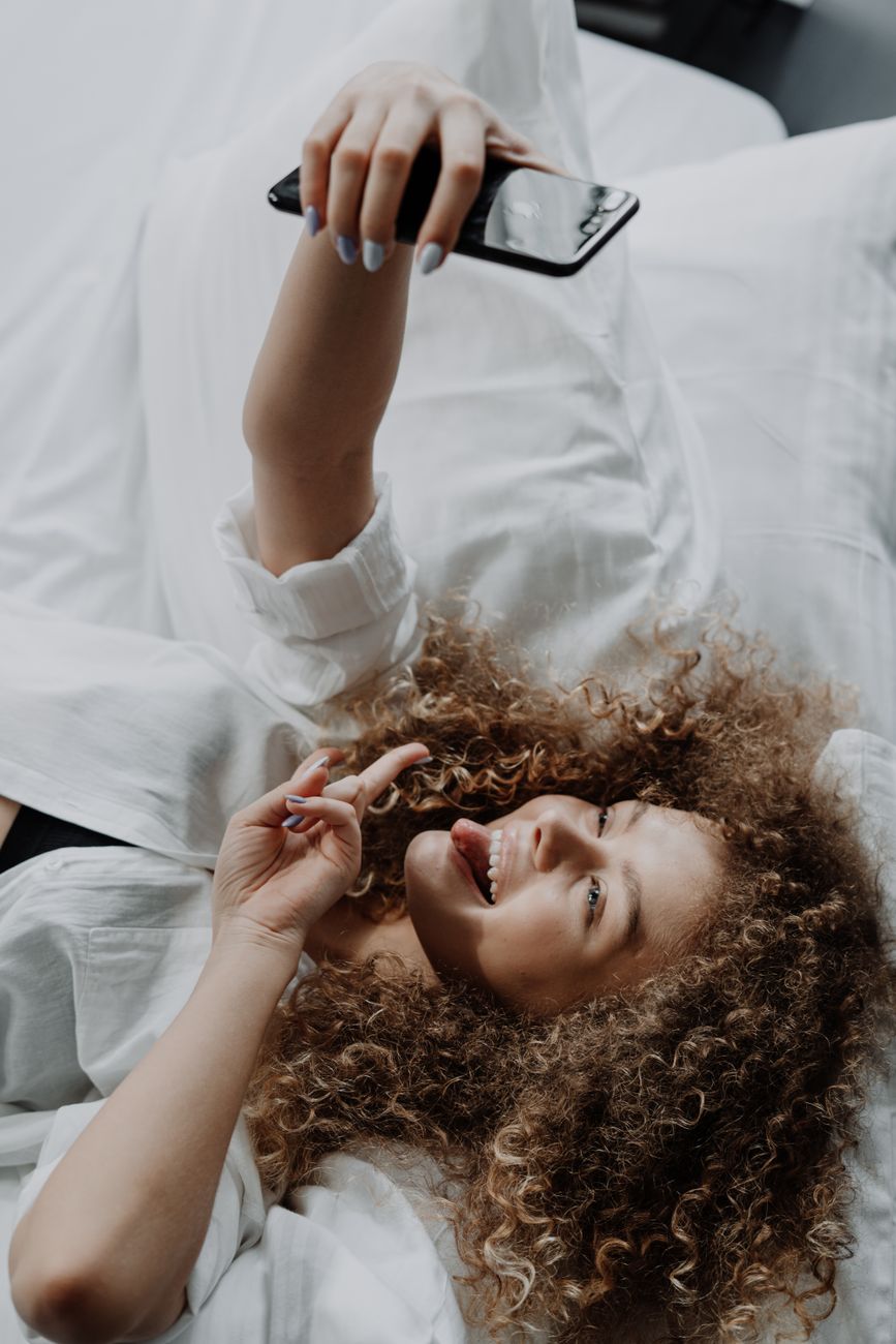 Smiling young woman with curly hair taking a selfie on a cozy bed, capturing a fun moment.