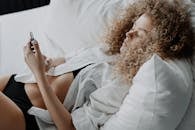 一名卷发女子躺在床上，穿着白衬衫打手机