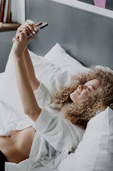 A woman with curly hair takes a selfie while lying on a bed in a bright, cozy bedroom.