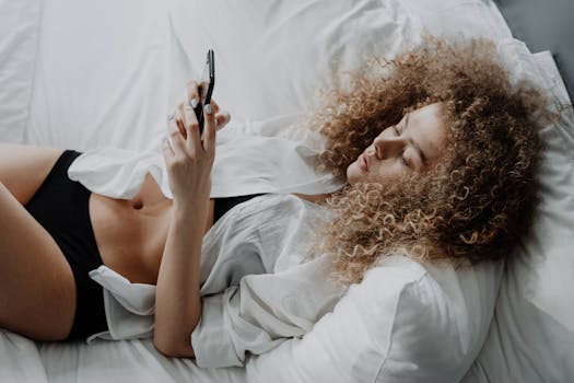 A woman with curly hair lies in bed, using a smartphone and relaxing on white linen.