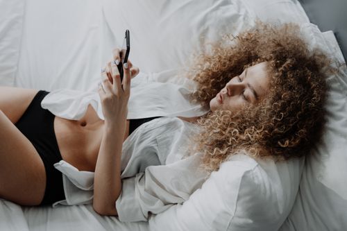 Free A woman with curly hair lies in bed, using a smartphone and relaxing on white linen. Stock Photo