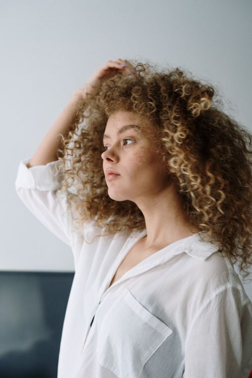 Free Young woman with vibrant curly hair wearing a white shirt, looking thoughtful indoors. Stock Photo