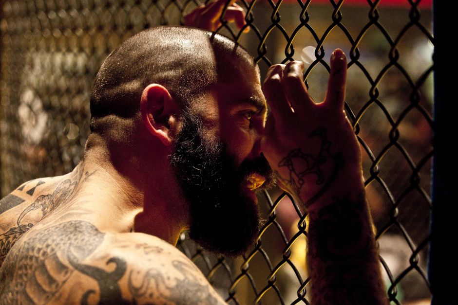 A bearded, tattooed man grips a chain-link fence, conveying intensity and solitude. A bearded, tattooed man grips a chain-link fence, conveying intensity and solitude.