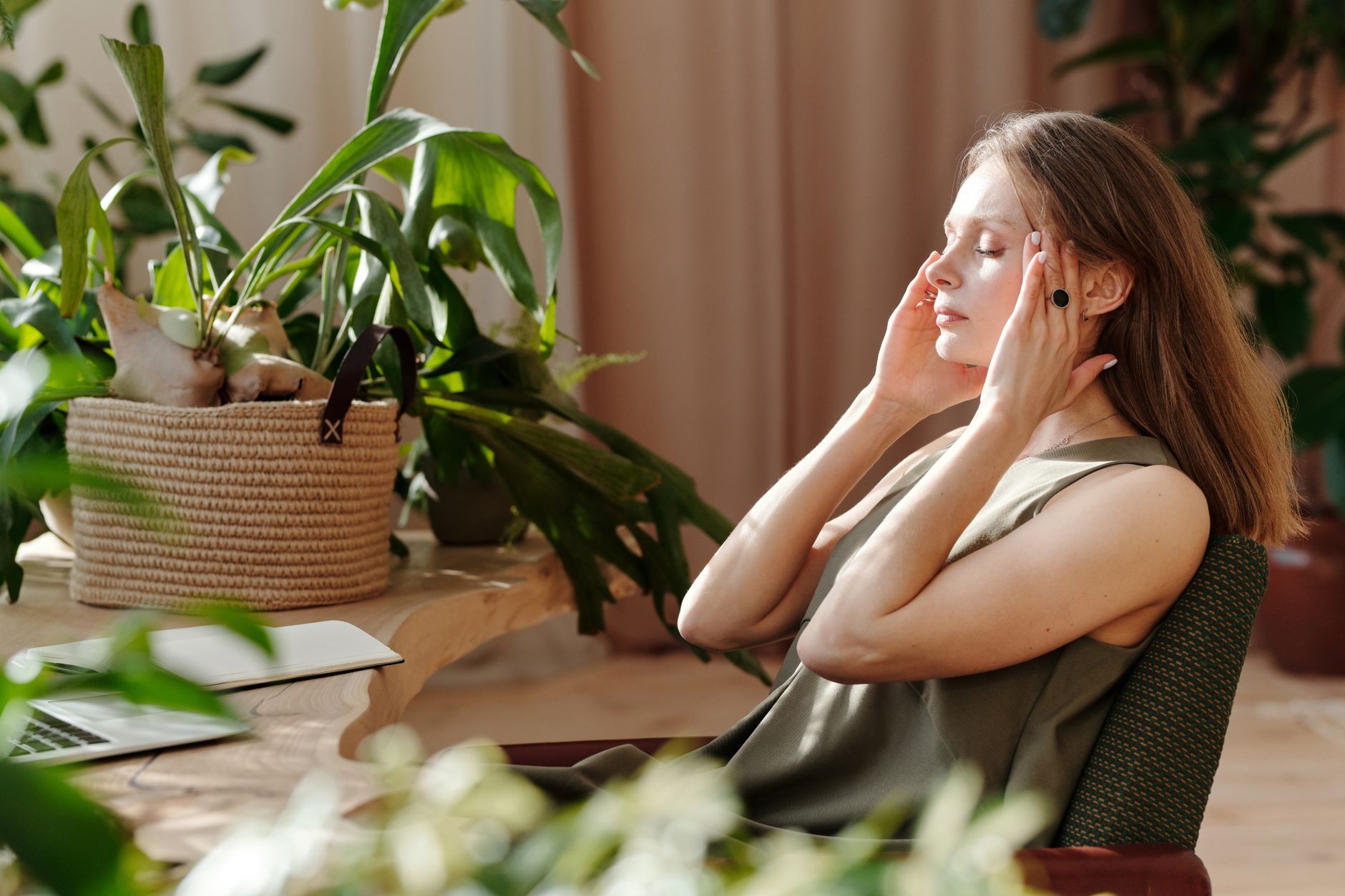 冥想教學中的「無努力」原則:定義、應用與益處 A woman relaxes, massaging her temples among indoor plants in a bright, sunlit room.