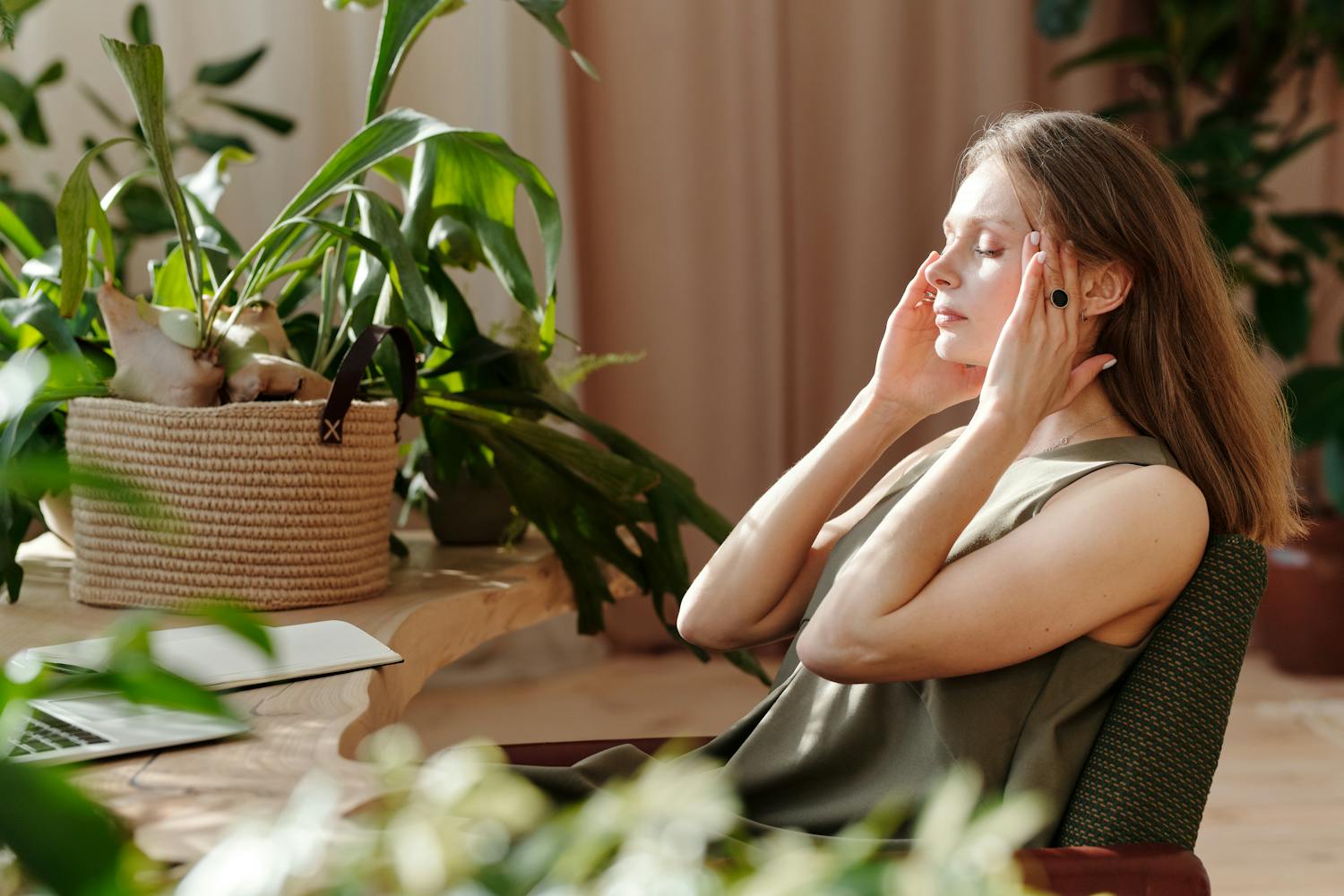 A woman relaxes, massaging her temples among indoor plants in a bright, sunlit room