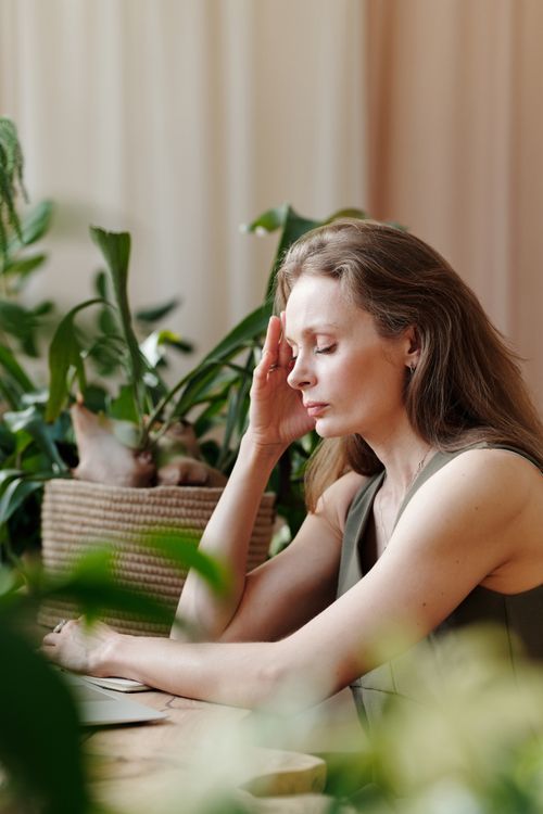 Free Adult woman sitting at home surrounded by plants, touching her temple with eyes closed, signifying a headache. Stock Photo