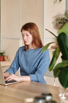 Caucasian woman in blue dress typing on a laptop at home office desk with plants.