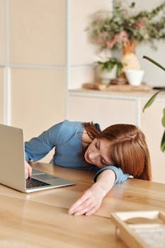 在现代化的办公室里，一个穿着蓝色连衣裙的女人躺在办公桌上睡着了，笔记本电脑就在旁边