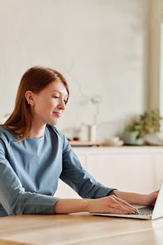 Smiling woman in blue shirt works on a laptop at home with a relaxed, bright atmosphere.