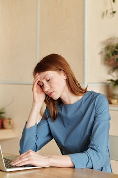 Exhausted woman in blue dress working at desk with laptop, conveying stress.