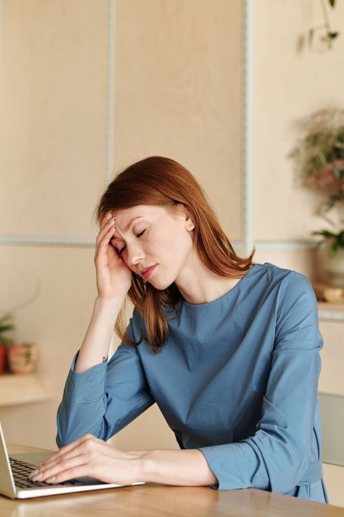 Free Exhausted woman in blue dress working at desk with laptop, conveying stress. Stock Photo