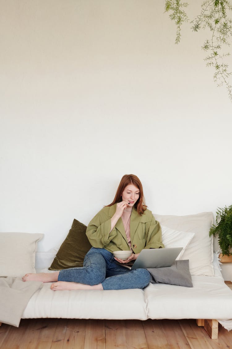 A Woman Sitting On A Couch Watching On A Laptop While Eating