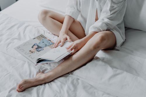 Free A young woman in a white shirt reads a magazine comfortably on a bed. Stock Photo