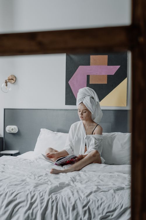 Free Young woman with a towel on head reading a book in a cozy, minimalist bedroom setting. Stock Photo