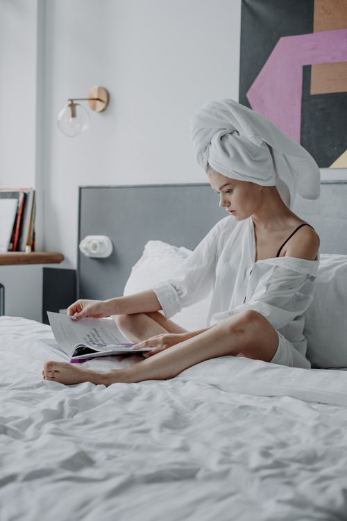 Free A young woman reading a magazine on a bed, wrapped in a towel, exuding relaxation and home comfort. Stock Photo