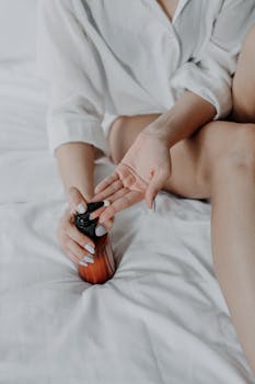Young woman applying lotion in cozy bedroom with white linens, embracing self-care routine.