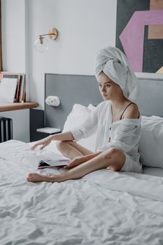 Young woman in a modern bedroom, wrapped in a towel, sitting and relaxing on a white bed.