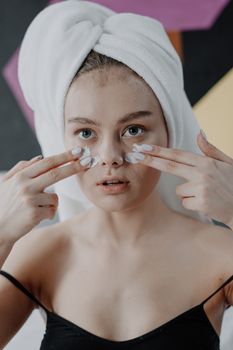 Portrait of a young woman applying cream with a towel wrapped on her head indoors.