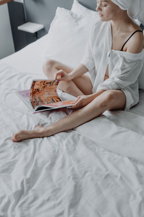 Free Young woman in a white shirt reads a magazine on a bed in a calm, natural light setting. Stock Photo