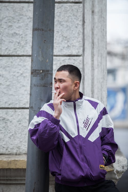 Young Asian man in sportive jacket with hand in pocket smoking cigarette at street post looking away