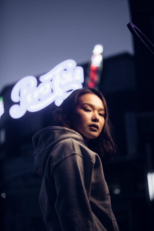 Low angle of serious Asian female standing near cafe entrance with neon sign in evening time and looking at camera